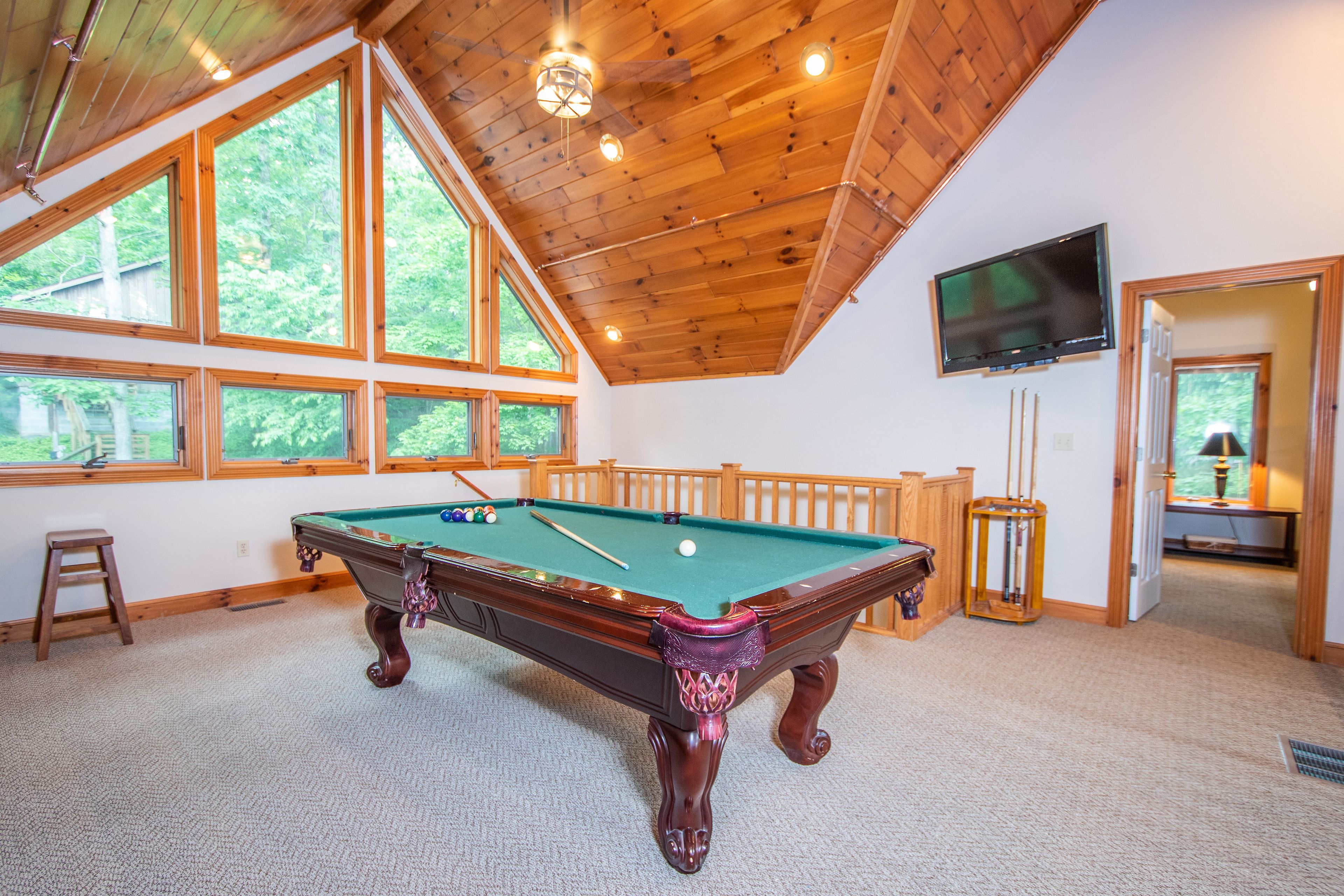 A pool table in a room with large windows, a television, and a wooden ceiling.