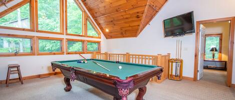 A pool table in a room with large windows, a television, and a wooden ceiling.
