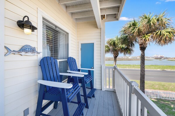 A porch with blue Adirondack chairs, a blue door, and a wall-mounted light fixture. - Relaxing Beach Escape With Balcony, Pool Access, and Short Walk to Shore (Corpus Christi)
