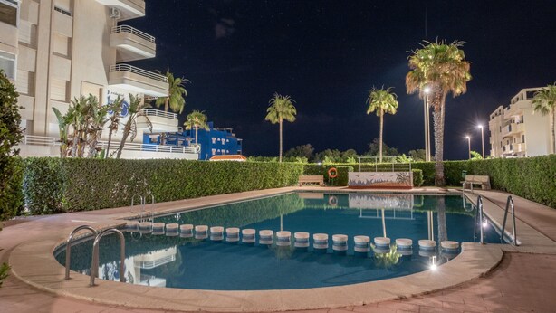A swimming pool with a surrounding hedge, palm trees, and a building with balconies. - Las Olas  4º Solo Familias! (Xeraco Playa)