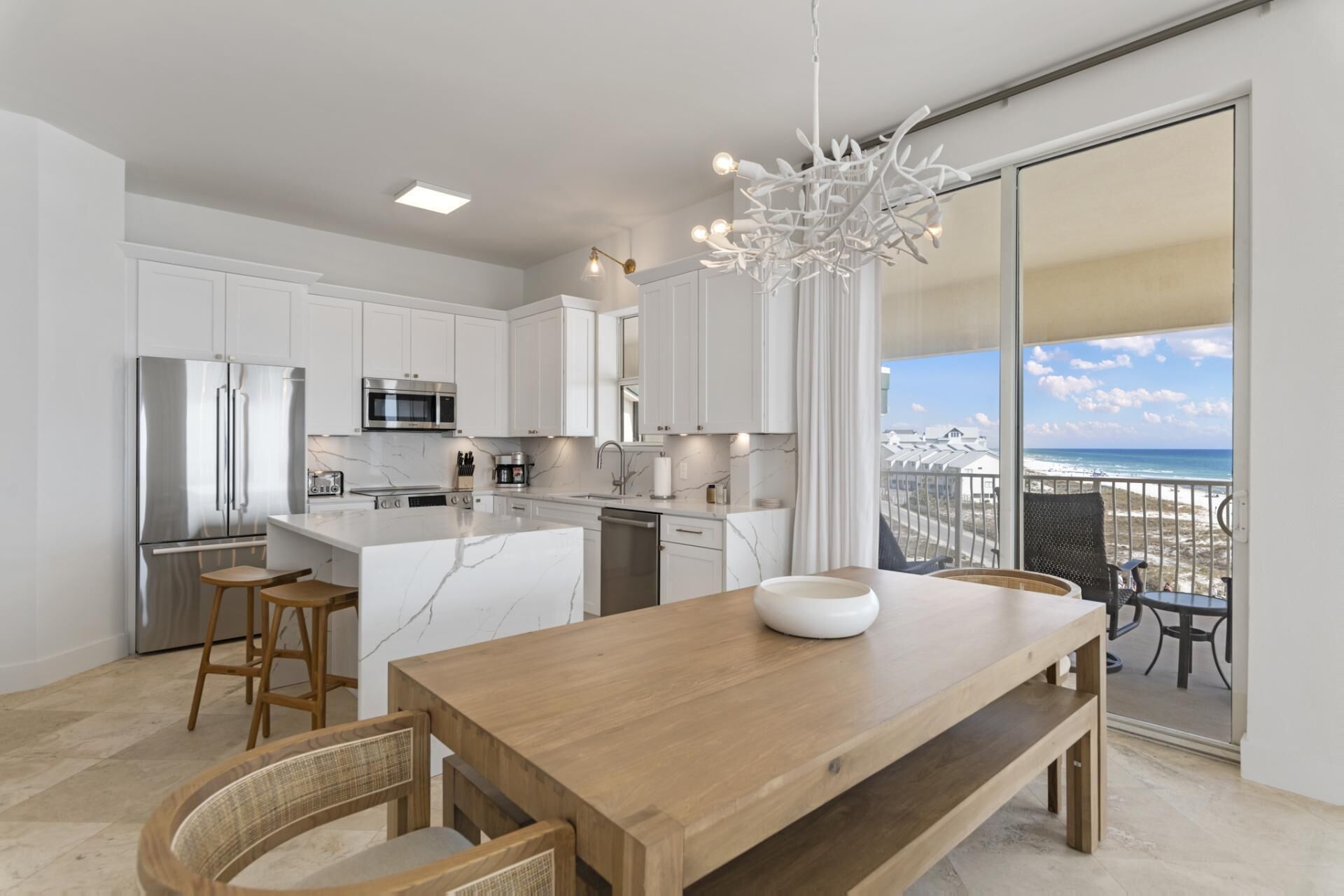 A modern kitchen with white cabinetry, stainless steel appliances, and a large island. There is a dining area with a wooden table and chairs, and a view of the beach through large sliding glass doors.
