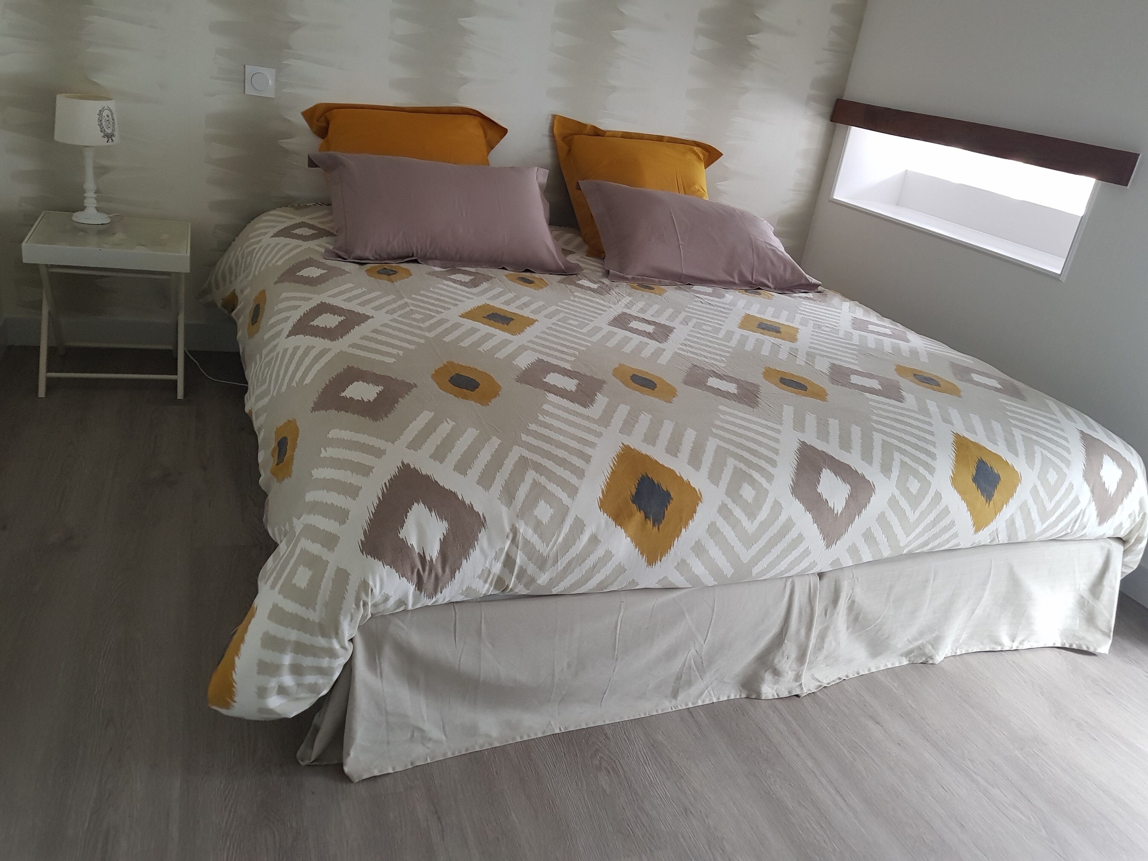 Una cama bien hecha con ropa de cama estampada, una mesita de noche con una lámpara y una ventana con vistas al cielo.