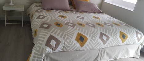 A neatly made bed with patterned bedding, a bedside table with a lamp, and a window with a view of the sky.