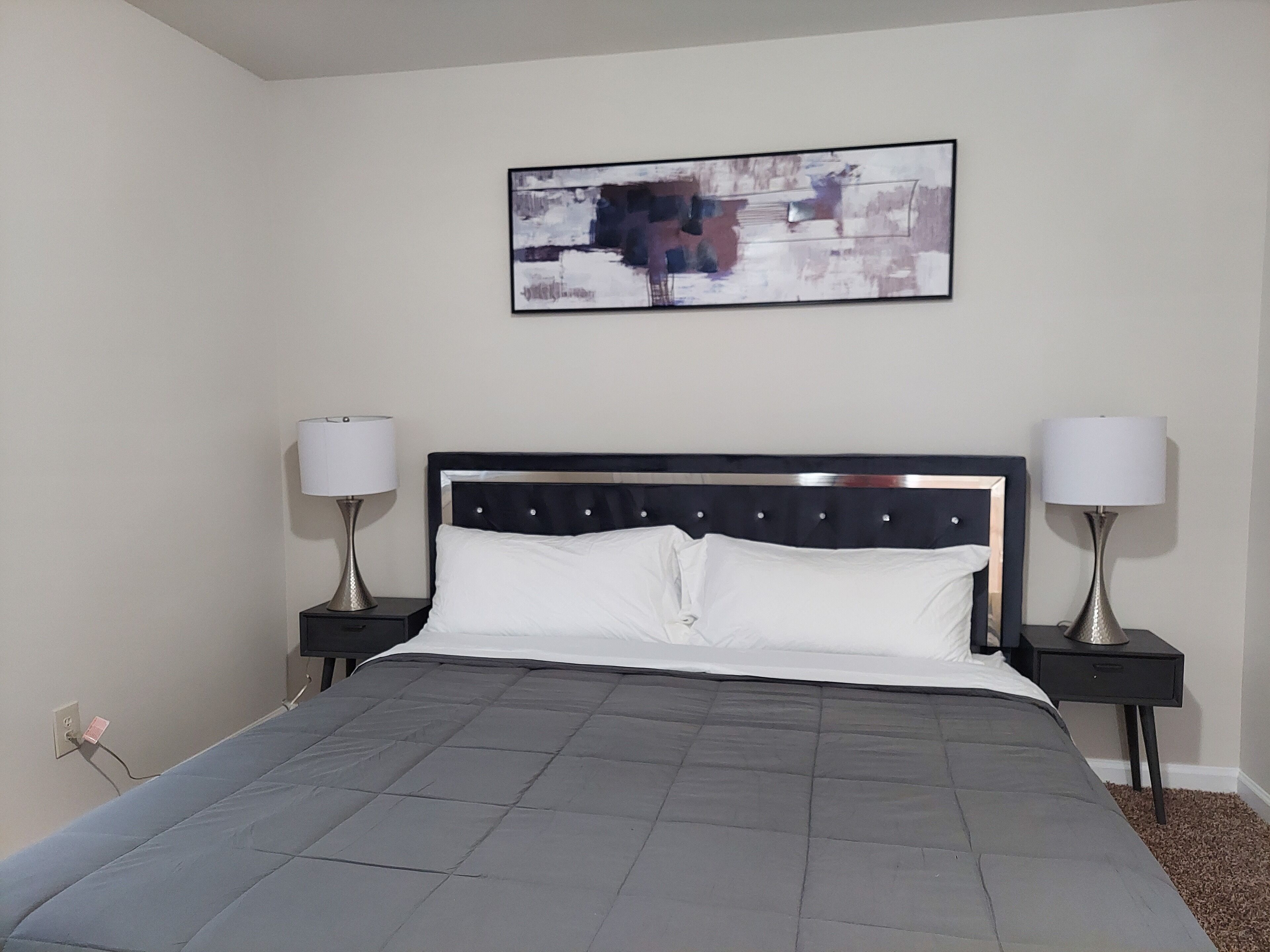Un lit bien fait, recouvert d’une couverture grise, deux tables de chevet avec des lampes, et un tableau abstrait encadré accroché au mur.