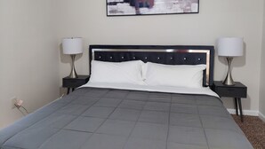 A neatly made bed with a grey quilt, two bedside tables with lamps, and a framed abstract painting on the wall.