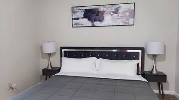 Un lit bien fait, recouvert d’une couverture grise, deux tables de chevet avec des lampes, et un tableau abstrait encadré accroché au mur.