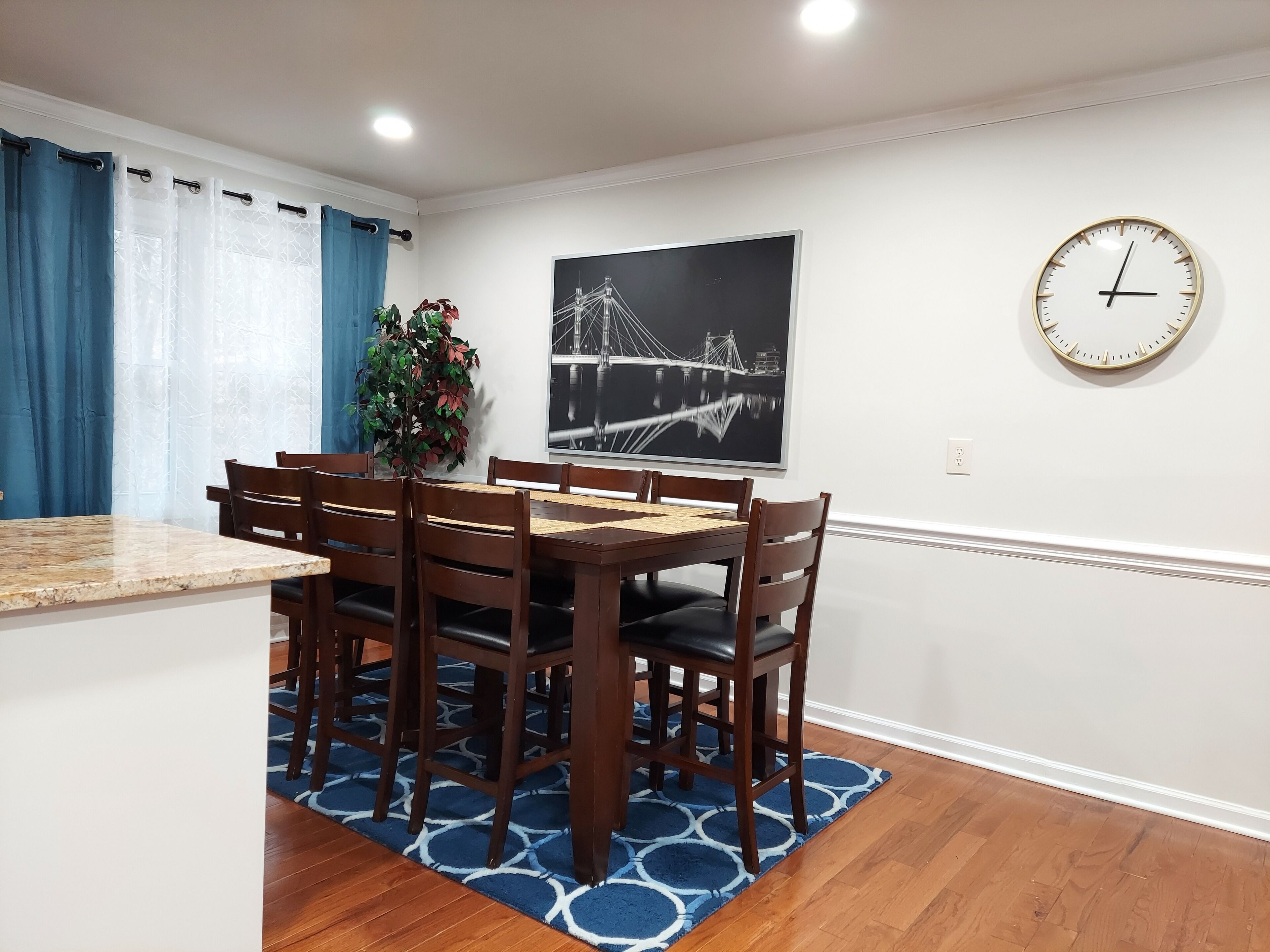 Un coin repas avec une table et des chaises en bois, un tapis à motifs bleus, une plante en pot, une horloge murale et un tableau encadré représentant un pont.