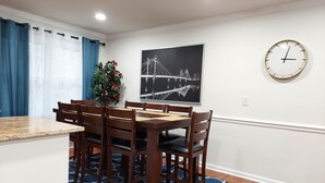 A dining area with a wooden table and chairs, a blue patterned rug, a potted plant, a wall-mounted clock, and a framed picture of a bridge.
