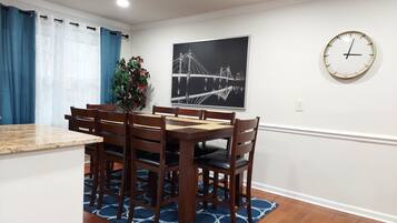 Un coin repas avec une table et des chaises en bois, un tapis à motifs bleus, une plante en pot, une horloge murale et un tableau encadré représentant un pont.
