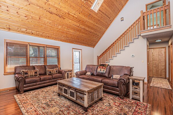 A spacious living room with two brown leather sofas, a wooden coffee table, and a large window with blinds. - Dancing Bear Lodge by Taylor Made Deep Creek (McHenry)