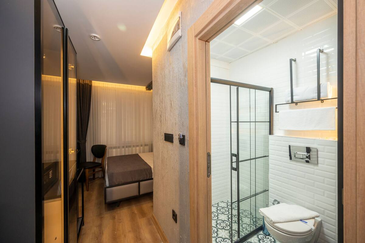 Una habitación compacta con una cama, una silla y una mesita. Junto a la habitación se encuentra un baño con cabina de ducha de cristal, un inodoro y un lavabo.