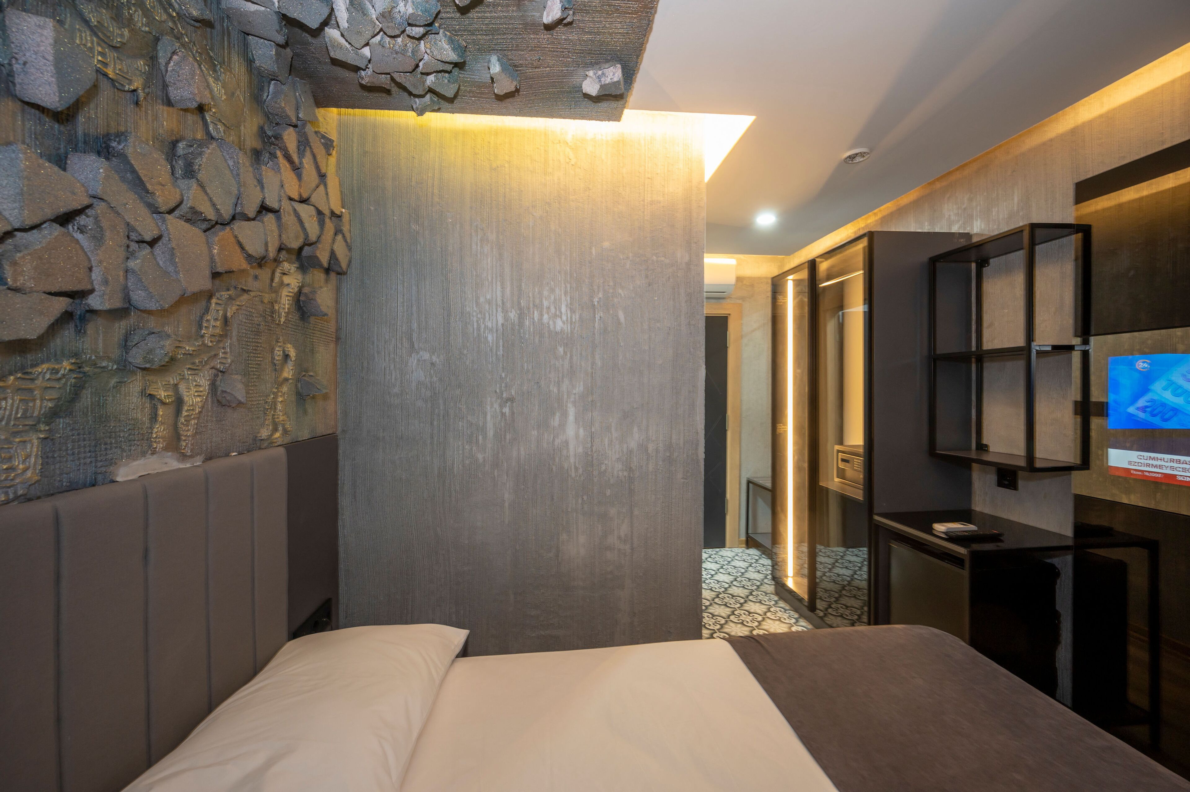 Een moderne hotelkamer met een stenen muur, een bed met beige beddengoed, een televisie aan de muur en een klein bureau.