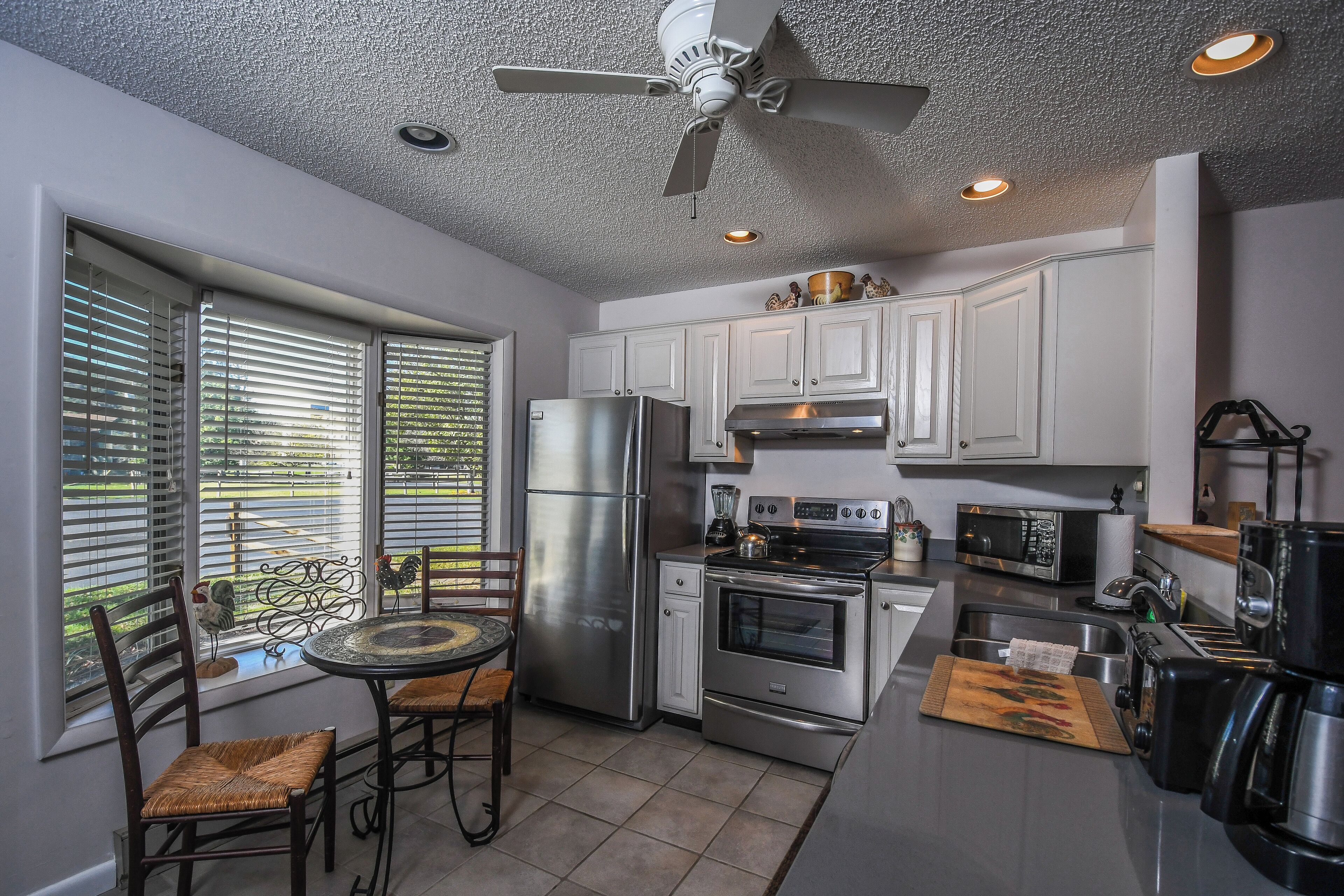 A modern kitchen with white cabinets, stainless steel appliances, a dining area with wicker chairs, and a ceiling fan.