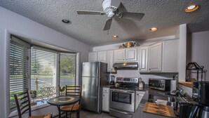 A modern kitchen with white cabinets, stainless steel appliances, a dining area with wicker chairs, and a ceiling fan. - Deep Creek Village #20 by Taylor Made Deep Creek (McHenry)