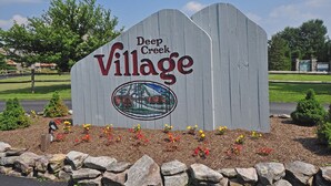 A wooden sign for Deep Creek Village with a logo and a landscaped area with flowers and rocks. - Deep Creek Village #29 by Taylor Made Deep Creek (McHenry)
