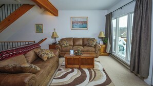 A living room with a brown leather sofa, a wooden coffee table, and a patterned rug. There is a painting on the wall and a window with curtains. - Deep Creek Village #29 by Taylor Made Deep Creek (McHenry)