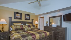 A bedroom with a large bed, two bedside lamps, a ceiling fan, a dresser, a mirror, and framed artwork on the wall. - Deep Creek Village #29 by Taylor Made Deep Creek (McHenry)
