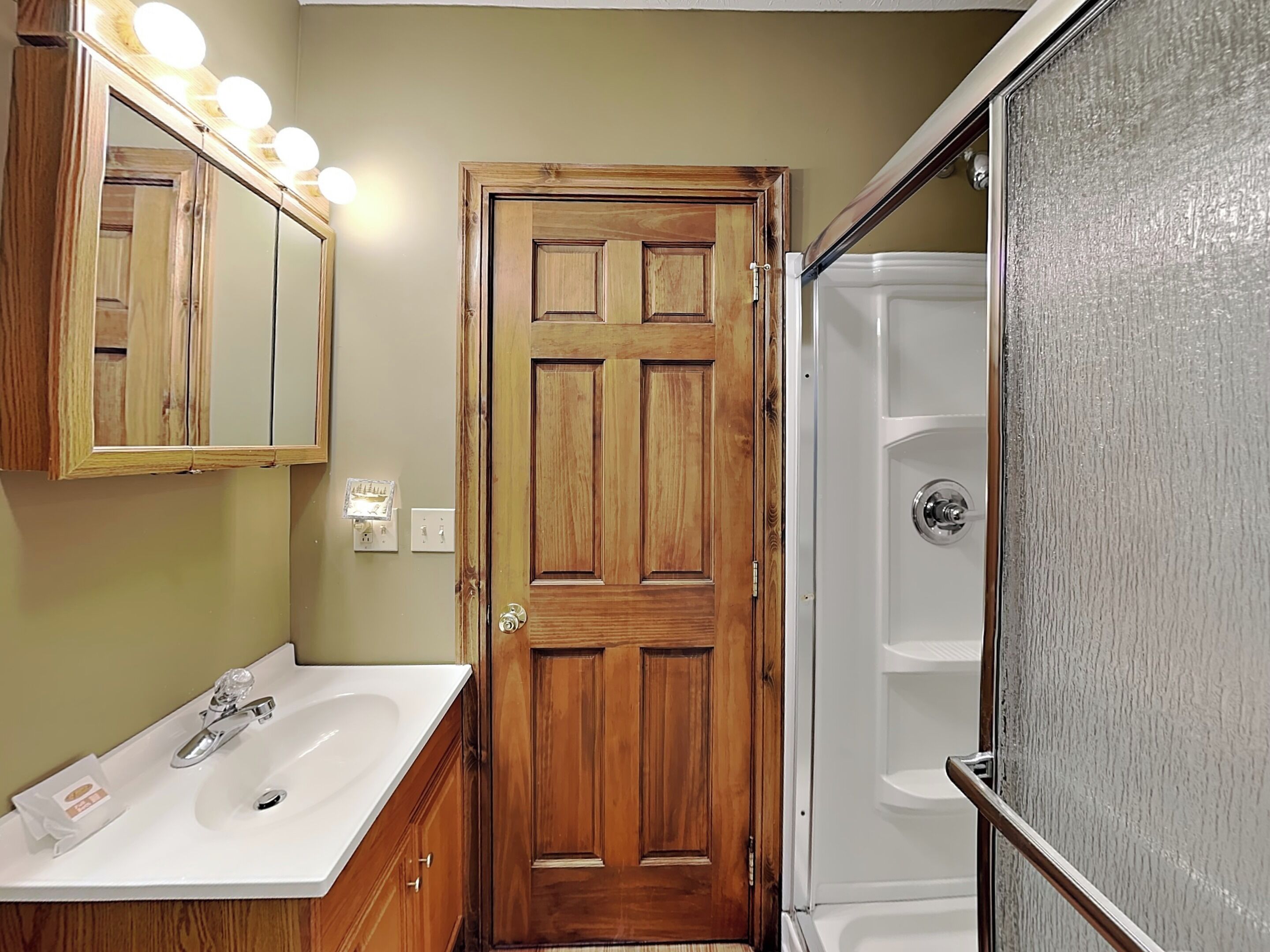 A bathroom with a wooden door, a white sink, a mirror with lights, and a glass shower enclosure.