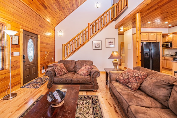 A cozy living room with a wooden staircase, leather sofas, a coffee table, and a kitchen area. - Donovan's Dacha by Taylor Made Deep Creek (McHenry)