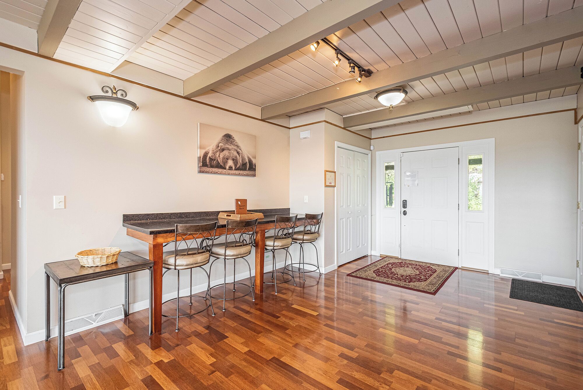 Una habitación amplia con piso de madera, una zona de bar con taburetes, una puerta blanca y un techo con vigas a la vista.