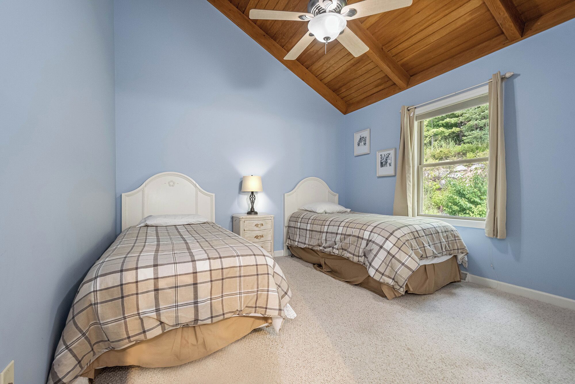 A bedroom with two beds, a ceiling fan, a window with a view of greenery, and a nightstand with a lamp.