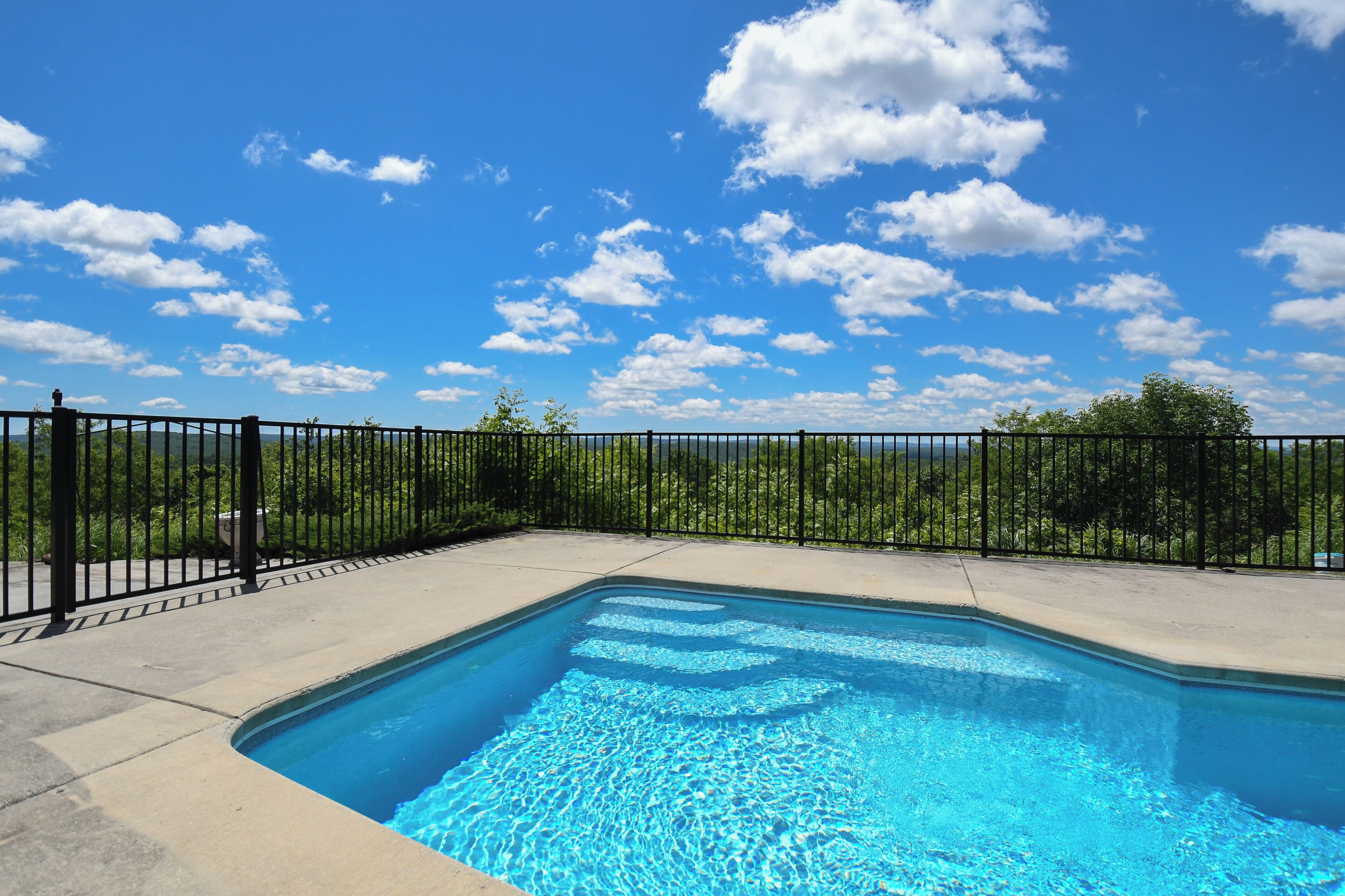 A rectangular swimming pool with a black metal fence, surrounded by a concrete deck, under a clear blue sky with scattered clouds.
