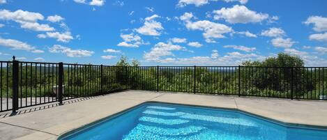 A rectangular swimming pool with a black metal fence, surrounded by a concrete deck, under a clear blue sky with scattered clouds.