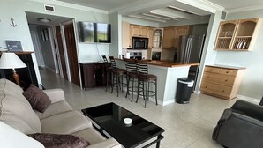A modern living room with a sofa, coffee table, and a patterned rug. A kitchen area with a microwave, stove, and bar stools is visible in the background. - Paradise in Pensacola Beach/Gulf of Mexico (Pensacola Beach)