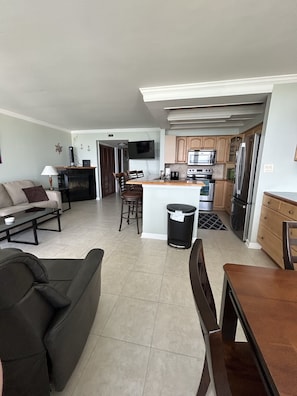 Uma sala de estar moderna com um sofá, mesa de centro e um espaço de jantar com mesa e cadeiras. Uma cozinha com eletrodomésticos e armários é visível ao fundo.