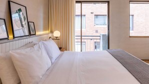 Ett hotellrum med en stor säng, ett fönster med utsikt över en byggnad med tegelstensfasad och inramade konstverk på väggen.