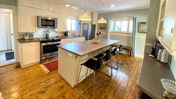 A modern kitchen with white cabinets, stainless steel appliances, a central island with bar stools, and wooden flooring.