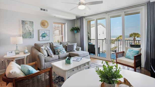 Una sala de estar moderna con un sofá gris, una mesa de centro blanca y sillas de mimbre. Hay un ventilador de techo, un ventanal con vista a la playa y una pequeña mesa con una lámpara y objetos decorativos.