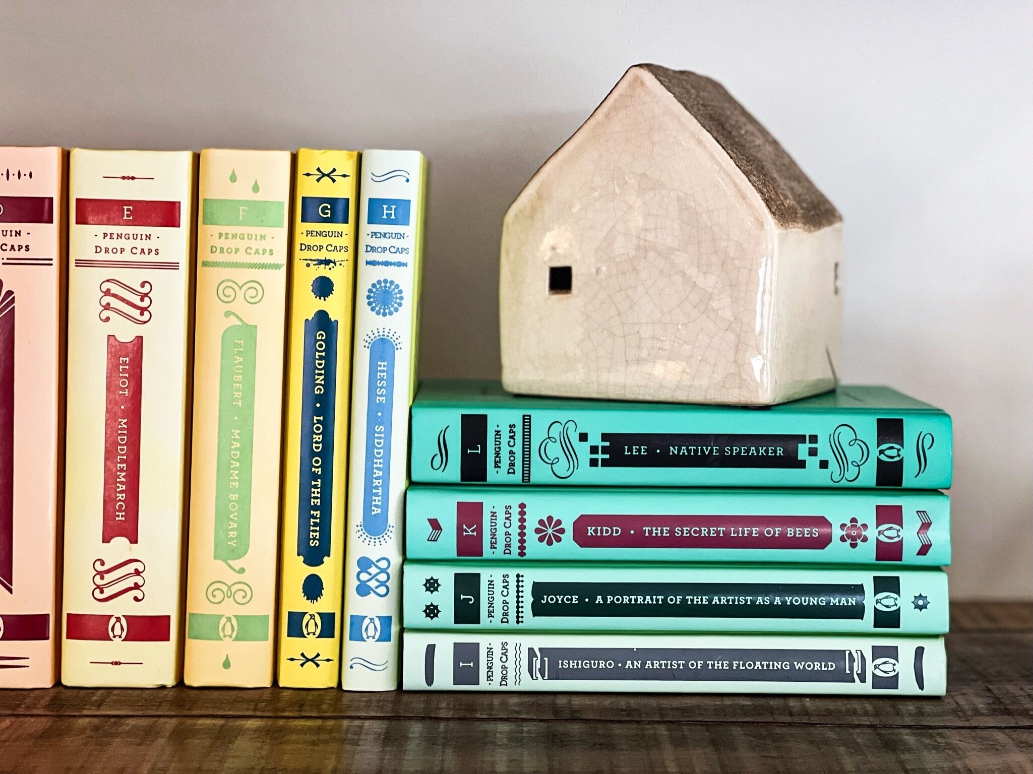 Eine Sammlung von Büchern mit unterschiedlichen Buchumschlägen, gestapelt auf einer Fläche, mit einem Keramik-Hausmodell obenauf.