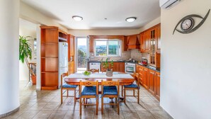 A kitchen with wooden cabinets, a dining table with chairs, and a view through the window. - Villa Wisteria - Three Bedroom Villa, Sleeps 6 (Latchi)