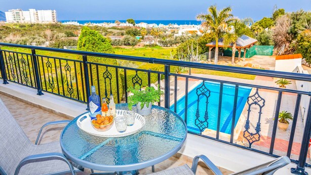 A balcony with a glass table set with drinks and snacks, overlooking a swimming pool and a coastal view. - Villa Wisteria - Three Bedroom Villa, Sleeps 6 (Latchi)