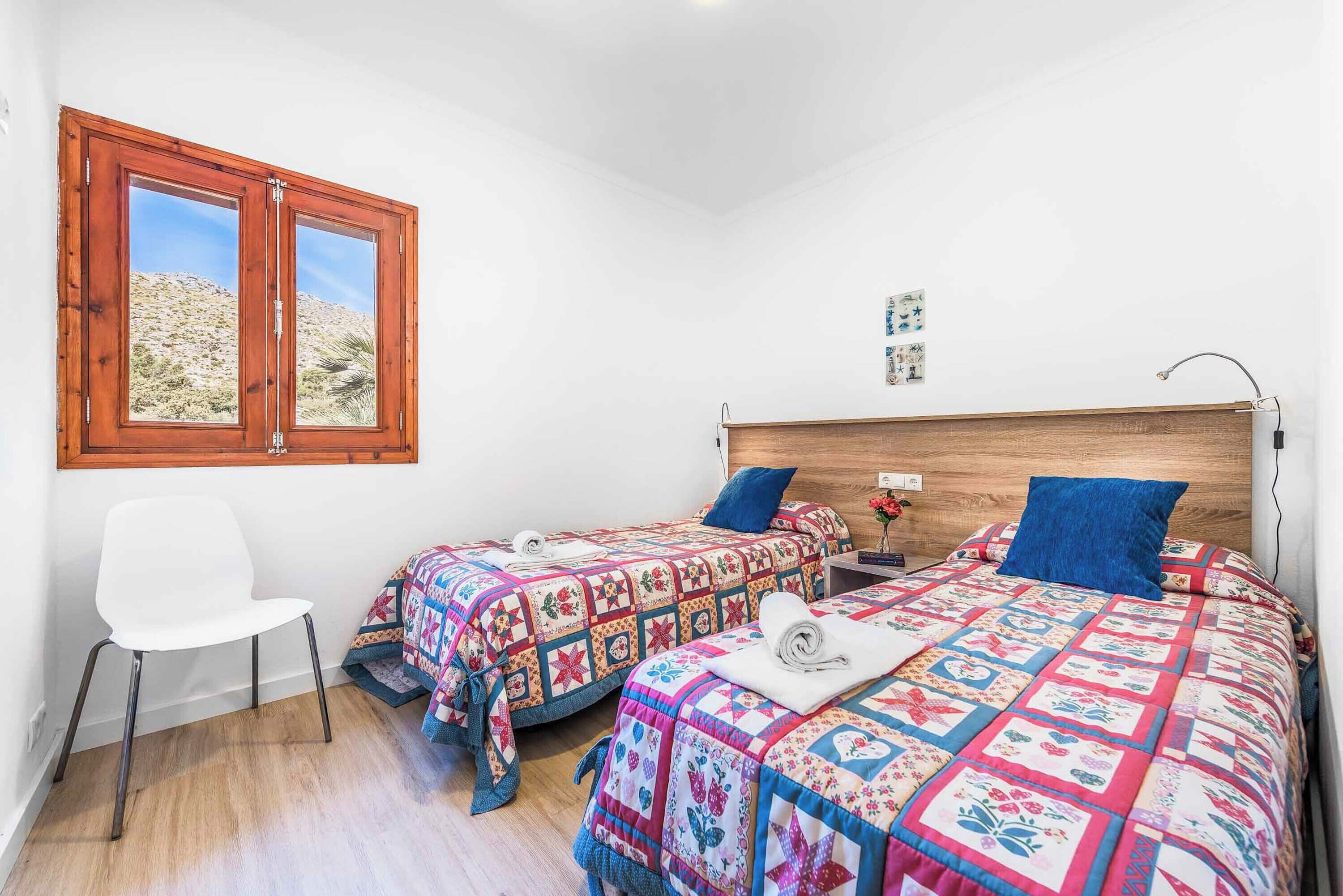 Ein Schlafzimmer mit zwei Betten, einem Holz-Kopfende, einem weißen Stuhl, einem Fenster mit Blick auf eine Wüste und einem an der Wand befestigten Nachttisch.