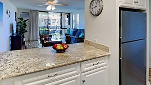 A kitchen with a granite countertop, a bowl of fruit, a clock, and a refrigerator. - here Elegance Meets the Breeze Kihei Renovated 2BR Condo Full Ocean Views (Kihei)