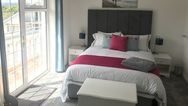 Ein modernes Schlafzimmer mit einem großen Bett, einem Nachttisch, einer Ablagefläche und Blick ins Freie durch eine Schiebetür.