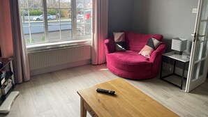 Ein Zimmer mit einem roten Sofa, einem Holztisch, einer Lampe und einem Fenster mit Blick auf geparkte Autos.