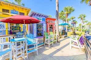 Um deck de madeira com cadeiras e guarda-sóis coloridos, cercado por palmeiras e lojas de praia.