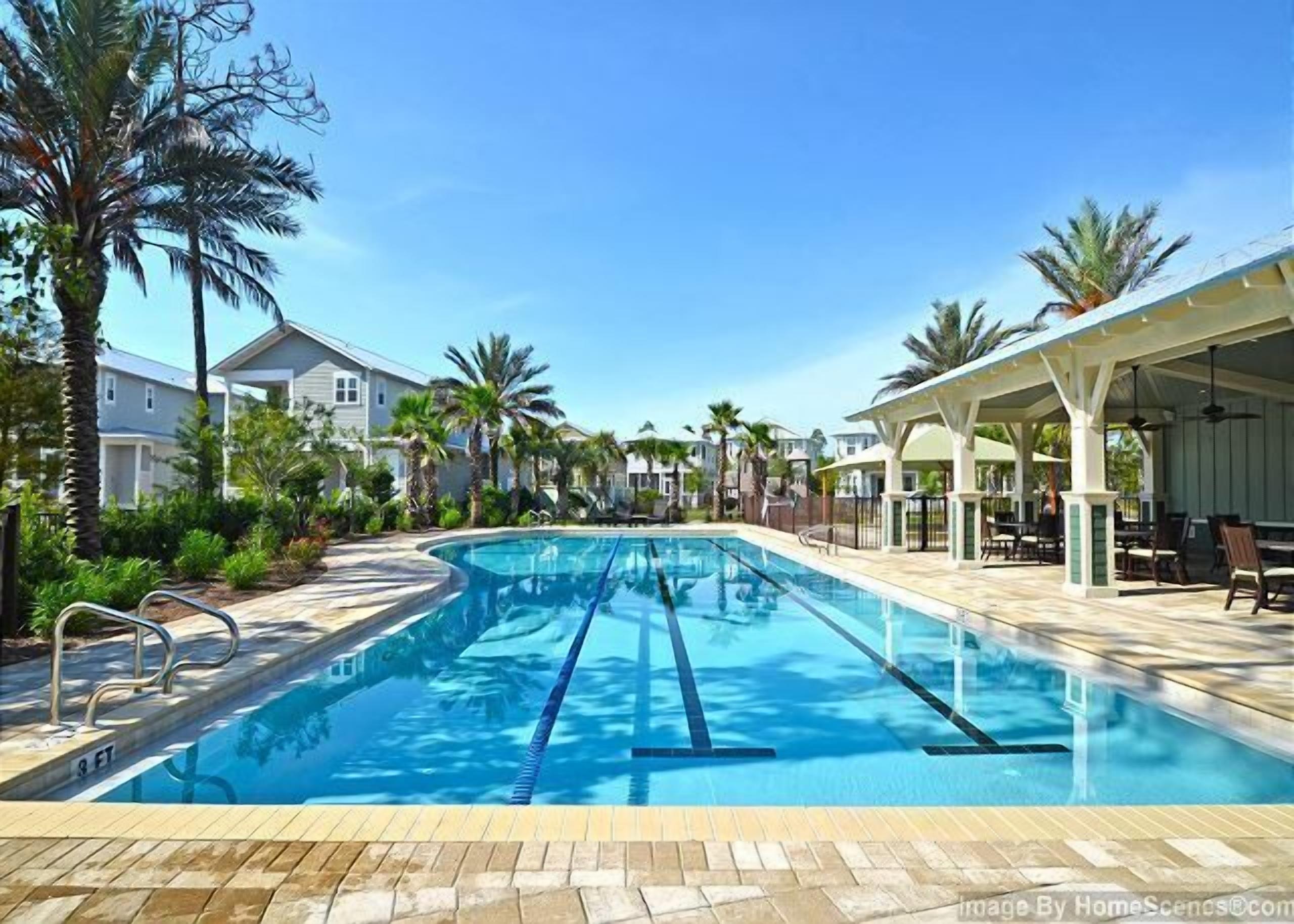A swimming pool with clear blue water, surrounded by palm trees and a covered patio area with outdoor seating.