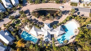 An aerial view of a resort with a central swimming pool, a circular water feature, and surrounding palm trees.