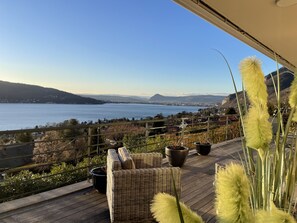 Ein Balkon mit Korbmöbeln, Blumentöpfen und Blick auf einen See sowie entfernte Berge.