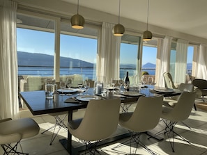 Een eetruimte met een tafel voor vier personen, grote ramen met uitzicht op zee en bergen, en moderne hanglampen.