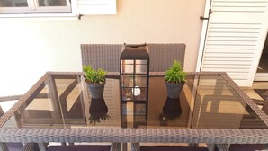 Une table à plateau en verre, agrémentée de deux plantes en pot, de chaises en osier et d’une lanterne.