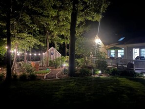 A residential property at night with a well-lit garden, a wooden fence, and a house with a porch. - Modern Family Getaway In Harwich (Harwich)