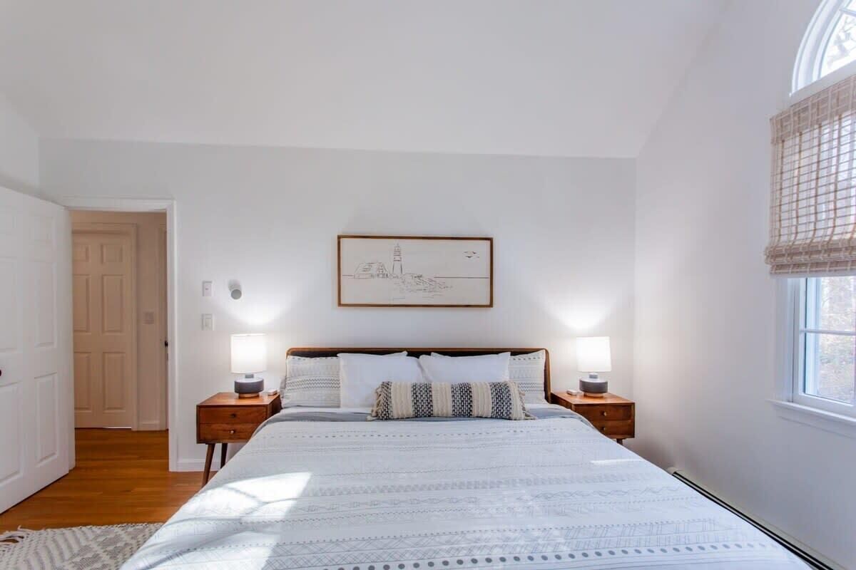 Une chambre à coucher avec un lit, deux tables de chevet avec des lampes, un tableau encadré au mur et une fenêtre avec un store.