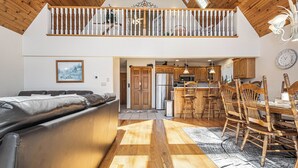 A spacious living area with a sofa, wooden flooring, a dining area with chairs, a kitchen with wooden cabinets, and a staircase leading to an upper level. - Dreamers Inn by Taylor Made Deep Creek (Swanton)