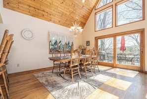 A dining area with a wooden table and chairs, a chandelier, and large windows.
