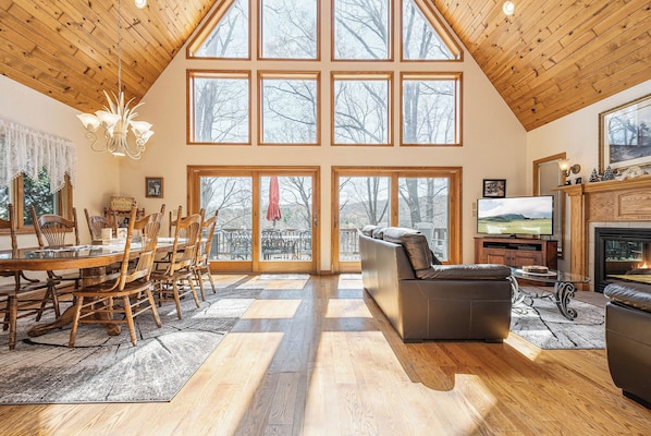 A spacious living room with a high ceiling, large windows, a fireplace, and a dining area with wooden furniture.